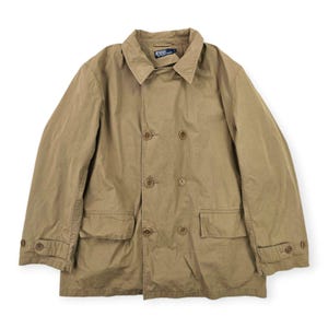 Op de afbeelding: Een beige dubbelrijige jas met een puntige kraag, lange mouwen met knoopmanchetten en twee voorzakken met klep. Het kledingstuk is voorzien van een Polo by Ralph Lauren-label bij de binnenhals en wordt gesloten met zes bruine knopen.