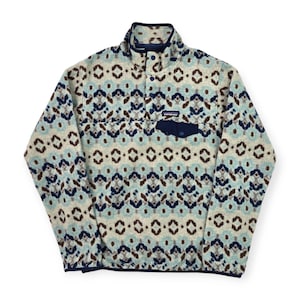 May include: A patterned fleece pullover in cream, light blue, and brown. The pullover features a high collar, a chest pocket, and a button-up closure. The design includes geometric shapes and the brand's logo.