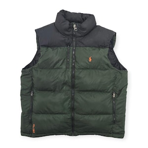 May include: A two-tone vest in forest green and black, featuring a high collar and a full-length zipper. The vest has a small orange logo on the chest and two zippered pockets. The vest is a puffy style, suitable for cold weather.