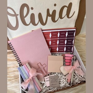 May include: A gift box with a pink notebook, Bible tabs, pens, highlighters, and a cross-shaped paper clip. The box includes a canvas bag with the name "Olivia" printed on it. The box also contains a small card with text.
