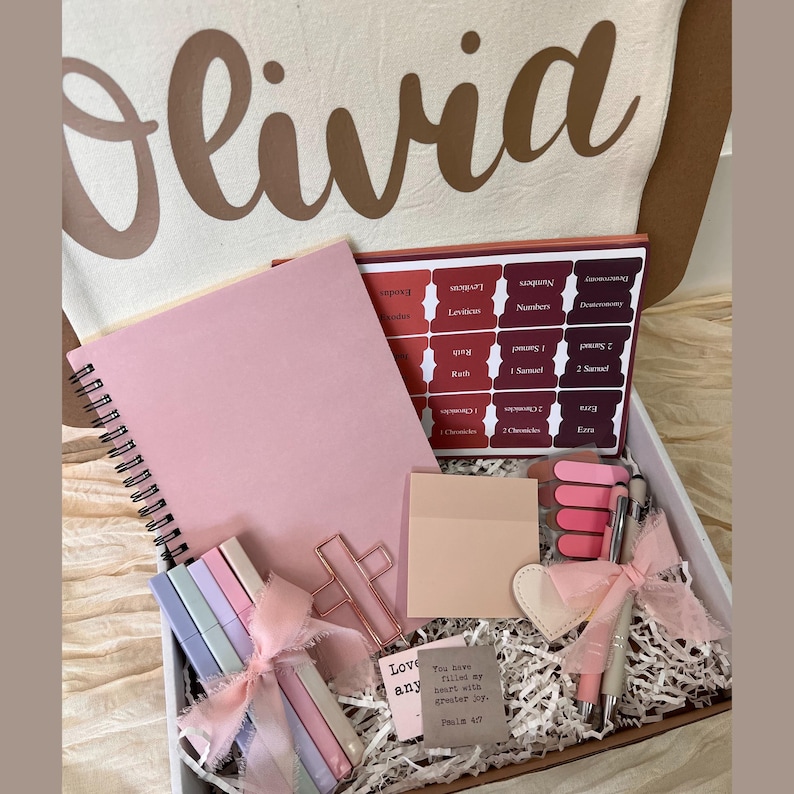 May include: A gift box with a pink notebook, Bible tabs, pens, highlighters, and a cross-shaped paper clip. The box includes a canvas bag with the name "Olivia" printed on it. The box also contains a small card with text.