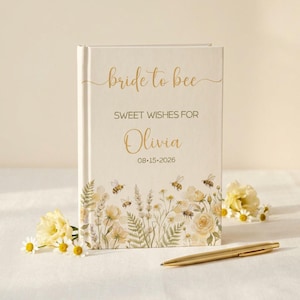 May include: A cream-colored hardcover book with the gold text bride to bee, SWEET WISHES FOR, Olivia, and 08.15.2026. The cover features a watercolor illustration of yellow flowers, green foliage, and small bees. A gold pen lies in front of the book.