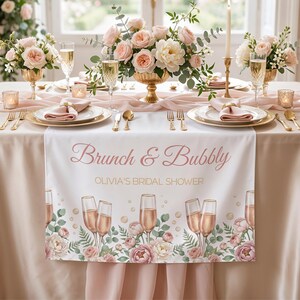 Pu&ograve; includere: Una tavola splendidamente apparecchiata per un addio al nubilato, con un runner bianco con la scritta "Brunch & Bubbly" e "Olivia's Bridal Shower". Il runner &egrave; decorato con calici di champagne e motivi floreali. La tavola &egrave; apparecchiata con posate, piatti e bicchieri dorati e ornata da centrotavola floreali.
