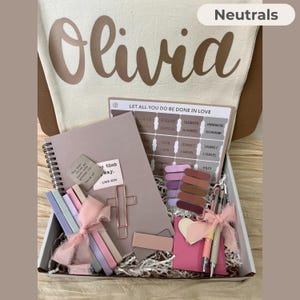May include: A gift box filled with neutral-toned stationery and accessories. Includes a notebook, pens, bookmarks, and a cross-shaped paper clip. The box also contains a tote bag with the name "Olivia" printed on it.