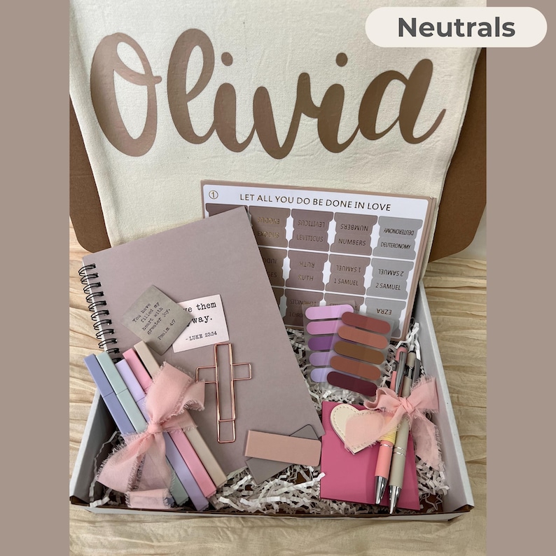 May include: A gift box filled with neutral-toned stationery and accessories. Includes a notebook, pens, bookmarks, and a cross-shaped paper clip. The box also contains a tote bag with the name "Olivia" printed on it.