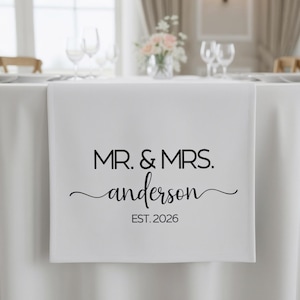 May include: White table runner with black text that reads "MR. & MRS. Anderson EST. 2026". The runner is draped over a white tablecloth, with a floral arrangement and glassware in the background.