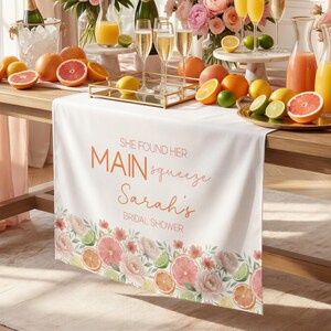 May include: White table runner with a floral and citrus fruit border. The runner has the text "SHE FOUND HER MAIN squeeze Sarah's BRIDAL SHOWER" in orange and pink. The table is set with drinks and fruit.