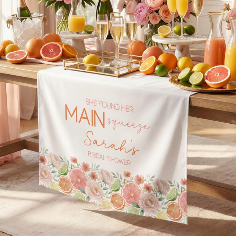 May include: White table runner with a floral and citrus fruit border. The runner has the text "SHE FOUND HER MAIN squeeze Sarah's BRIDAL SHOWER" in orange and pink. The table is set with drinks and fruit.