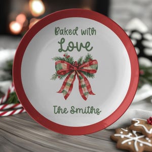 May include: A white ceramic plate with a red rim. The plate features the text "Baked with Love" and "The Smiths" in green script. A watercolor illustration of a red and green plaid bow with greenery is in the center.