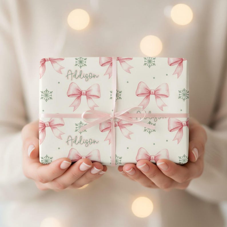 A person holds a horizontally oriented rectangular gift wrapped in cream paper with pink bows and green snowflakes, tied with pink ribbon.