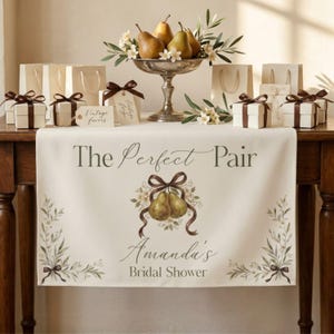 Puede incluir: Un camino de mesa color crema con el texto The Perfect Pair y Amanda's Bridal Shower, decorado con una ilustración de peras atadas con una cinta marrón y motivos de ramas de olivo, expuesto sobre una mesa con cajas de regalo.