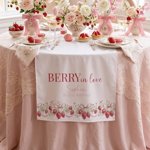 May include: A table set for a bridal shower, featuring pink roses, strawberries, and desserts. A white table runner with the text "BERRY in love Sophia's BRIDAL SHOWER" is the centerpiece. The table is decorated with pink accents.