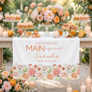 May include: A bridal shower table setting with a white table runner featuring the text "She Found Her Main Squeeze Sarah's Bridal Shower." The table is decorated with cupcakes, macarons, flowers, candles, and citrus fruits.