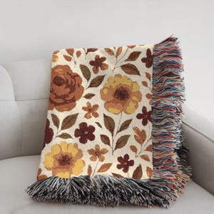 May include: A floral throw blanket with a cream background and a pattern of brown, yellow, and burgundy flowers and leaves. The blanket has a fringed edge in shades of black, white, and other colors. The blanket is folded over a white sofa.