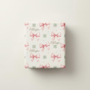 A flat view of a rectangular sheet of cream wrapping paper patterned with pink bows, green snowflakes, and personalized text.