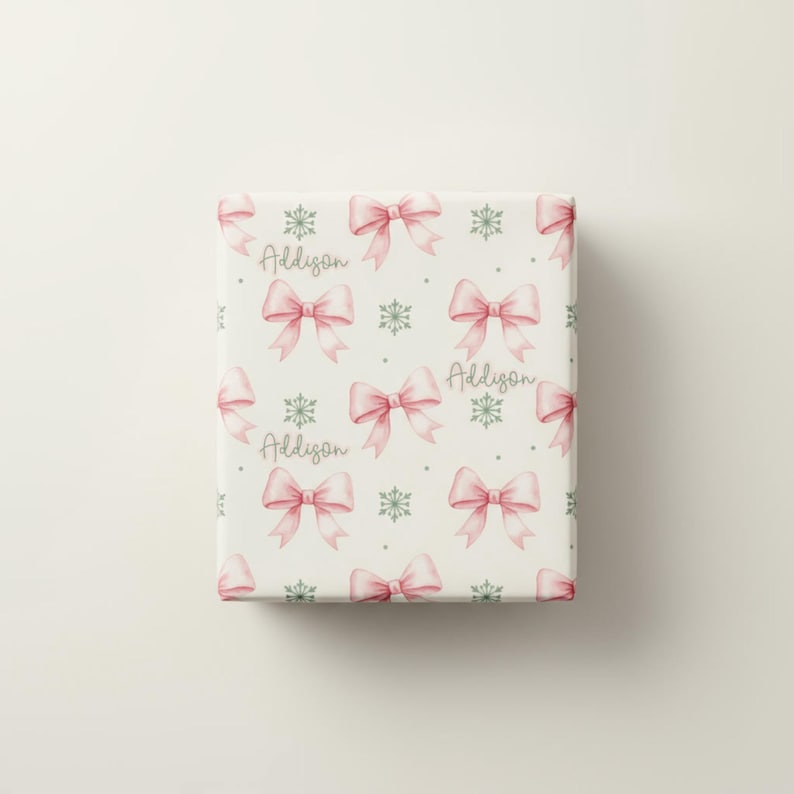 A flat view of a rectangular sheet of cream wrapping paper patterned with pink bows, green snowflakes, and personalized text.