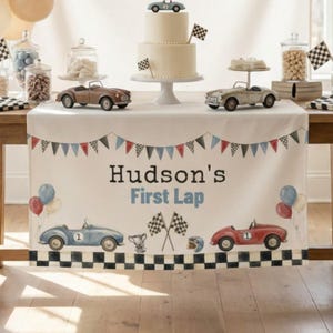 以下が含まれることがあります： レースをテーマにした白いテーブルランナー。バナー、"Hudson's First Lap"の文字、青と赤のヴィンテージ風レーシングカー、チェッカーフラッグ、トロフィー、ヘルメット、風船が描かれ、縁はチェッカー模様で仕上げられています。