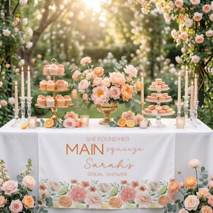 May include: A beautifully decorated table for a bridal shower, featuring cupcakes, macarons, and floral arrangements in peach and cream tones. The table has a white tablecloth with a banner that reads "She Found Her Main Squeeze Sarah's Bridal Shower."