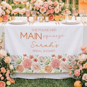 May include: A white table runner with the text "SHE FOUND HER MAIN squeeze Sarah's BRIDAL SHOWER" in copper-colored script. The runner is decorated with floral and citrus fruit illustrations, including oranges, grapefruits, and limes. The table is set for a bridal shower.