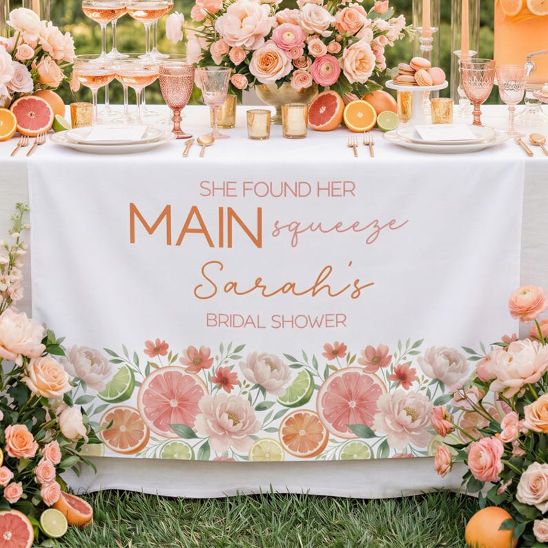 May include: A white table runner with the text "SHE FOUND HER MAIN squeeze Sarah's BRIDAL SHOWER" in copper-colored script. The runner is decorated with floral and citrus fruit illustrations, including oranges, grapefruits, and limes. The table is set for a bridal shower.