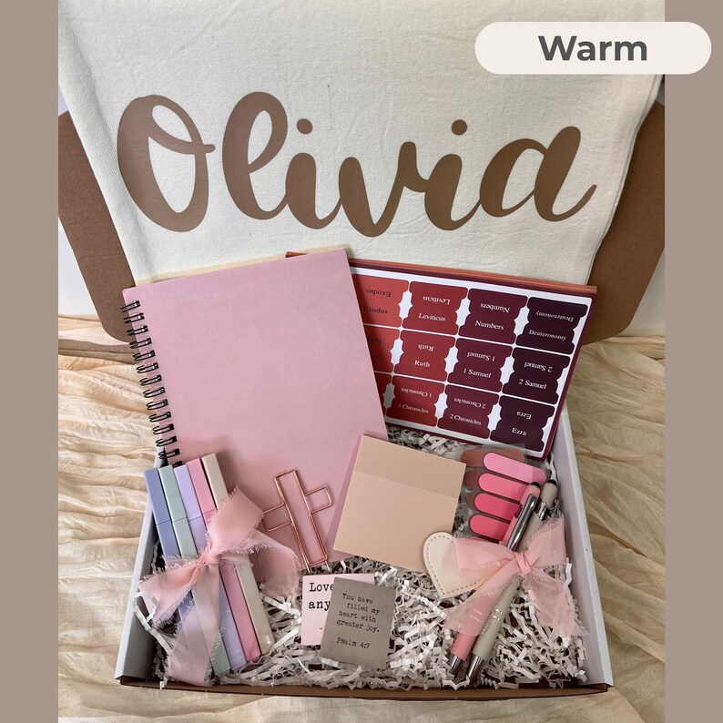 May include: A gift box filled with stationery items, including a pink notebook, pens, and a set of colorful clips. The box also contains a decorative cross-shaped paper clip and a small card with text. The box is lined with white paper.