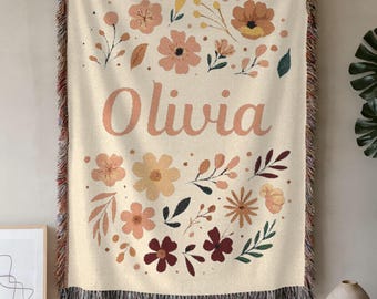 May include: Cream-colored woven throw blanket with a floral design and the name "Olivia" in peach-colored script. The blanket features a fringe border and a pattern of peach, yellow, and burgundy flowers and leaves.