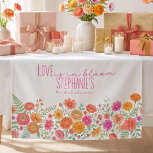 May include: A white table runner with a floral border in shades of pink, orange, and yellow. The text "LOVE is in bloom STEPHANIE'S bridal shower" is printed in pink. The table is set with gifts, candles, and flowers.