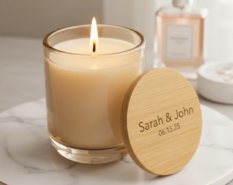 Puede incluir: Una vela encendida en un tarro de cristal transparente con tapa de madera. La tapa está grabada con "Sarah & John" y "06.15.25". La vela está sobre una bandeja de mármol blanco.
