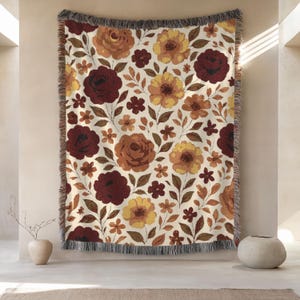 May include: A woven throw blanket with a floral pattern. The design features large burgundy, yellow, and orange flowers with green leaves on a cream background. The blanket has a fringed edge.