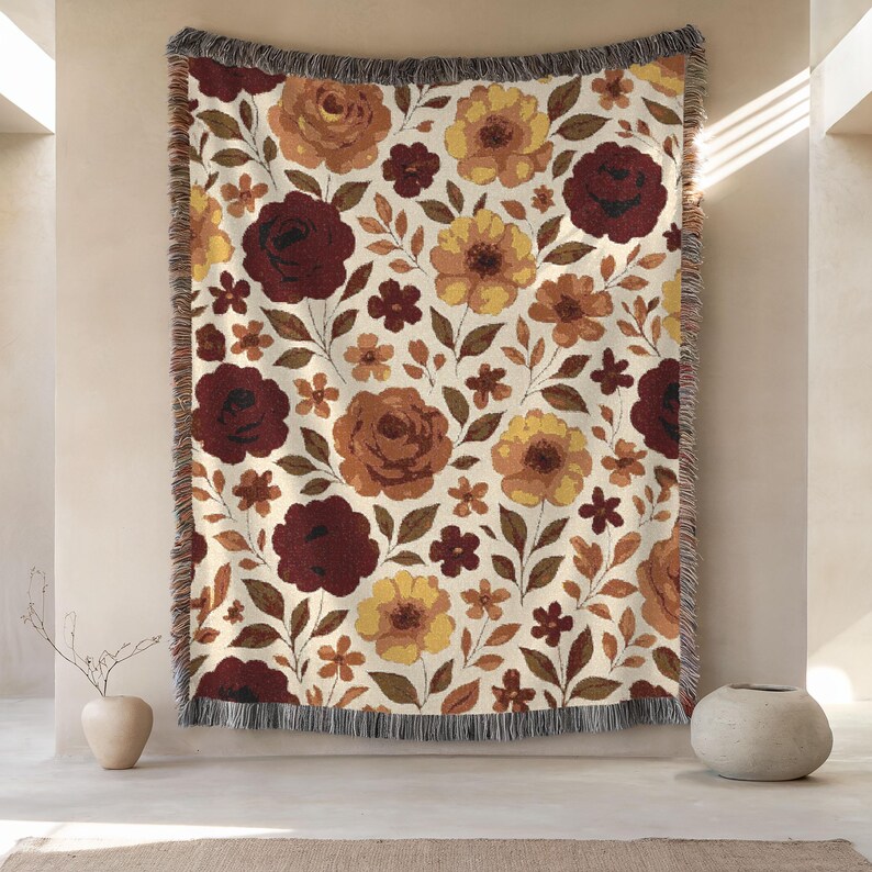 May include: A woven throw blanket with a floral pattern. The design features large burgundy, yellow, and orange flowers with green leaves on a cream background. The blanket has a fringed edge.