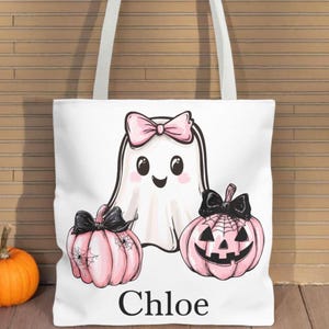 May include: White tote bag featuring a cartoon ghost with a pink bow, two pink pumpkins with black bows, and the name "Chloe" in black. The pumpkins have spiderwebs and jack-o'-lantern faces. The bag has white straps and is perfect for Halloween.