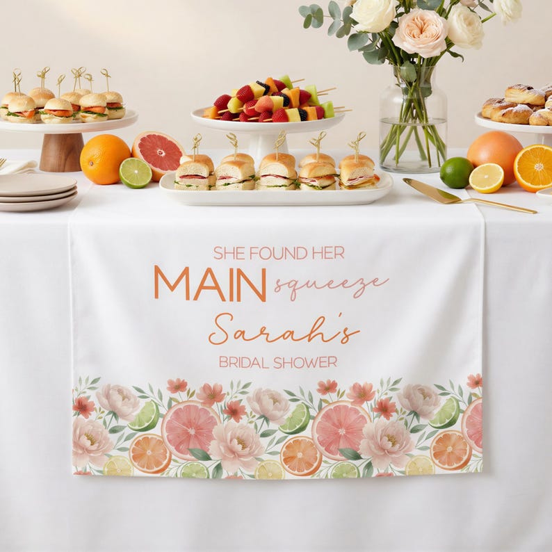 May include: White table runner with floral and citrus fruit border. Text reads "SHE FOUND HER MAIN squeeze Sarah's BRIDAL SHOWER" in orange script. The table is set with food and flowers.
