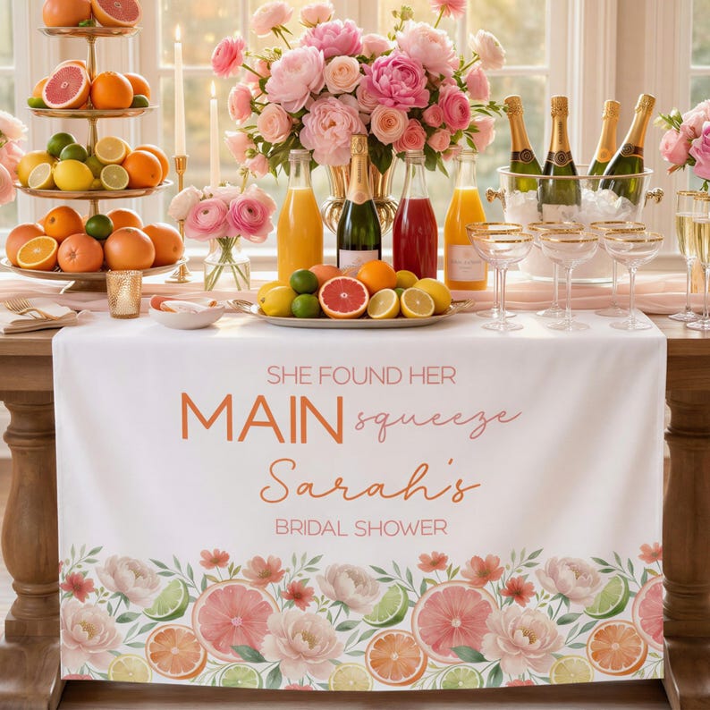 May include: A white table runner with the text "SHE FOUND HER MAIN squeeze Sarah's BRIDAL SHOWER" in orange script. The runner has a floral and citrus fruit border. Champagne, juice, and fruit are on the table.
