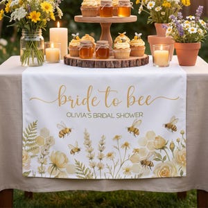 Puede incluir: Camino de mesa blanco con el texto dorado "bride to bee" y "Olivia's bridal shower". El camino de mesa está decorado con abejas, flores y panales de acuarela. Cupcakes, tarros de miel y velas sobre la mesa.