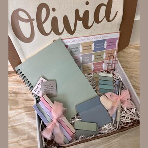 May include: A gift box with a personalized canvas bag, a spiral notebook, pens, sticky notes, and a card with the text "LET ALL YOU DO BE DONE IN LOVE". The box contains decorative ribbons and paper filler.
