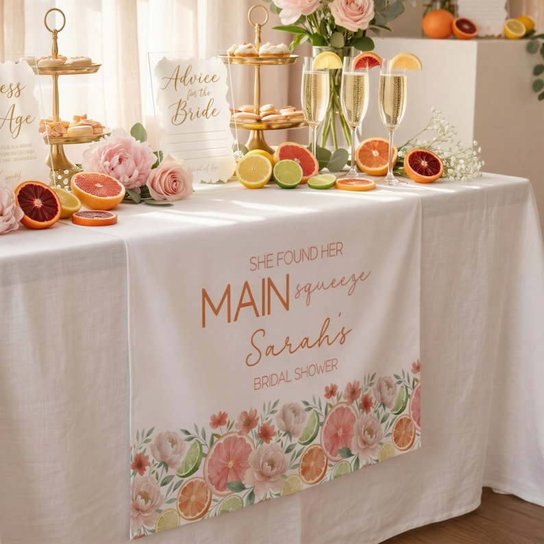 May include: A white table runner with the text "She Found Her Main Squeeze Sarah's Bridal Shower" in copper-colored script. The runner features a floral and citrus fruit border, with pink roses and sliced citrus fruits.