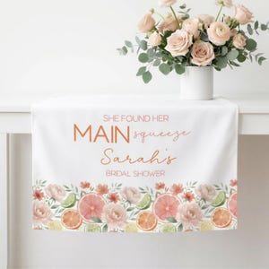May include: White table runner with floral and citrus fruit border. Text reads "SHE FOUND HER MAIN squeeze Sarah's BRIDAL SHOWER". A bouquet of pink roses in a white vase sits on the table.
