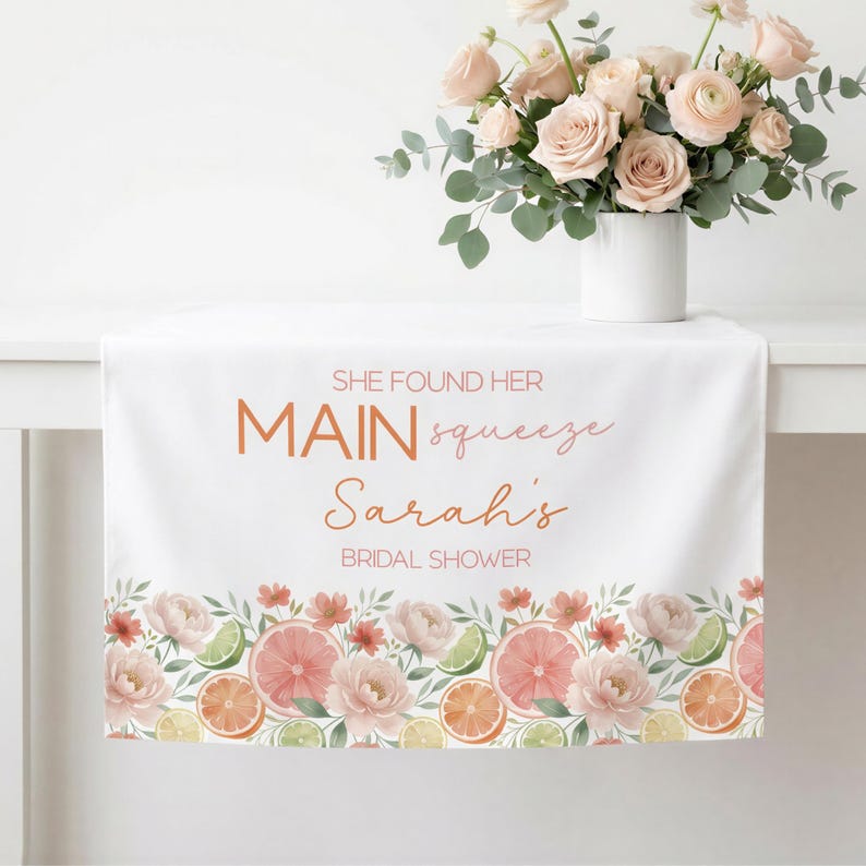 May include: White table runner with floral and citrus fruit border. Text reads "SHE FOUND HER MAIN squeeze Sarah's BRIDAL SHOWER". A bouquet of pink roses in a white vase sits on the table.