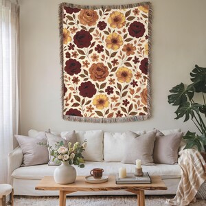 May include: A floral tapestry with a cream background and a repeating pattern of burgundy, brown, and yellow flowers. The tapestry is rectangular with a fringed edge and is hanging on a wall. The room has a white sofa, wooden coffee table, and decorative pillows.