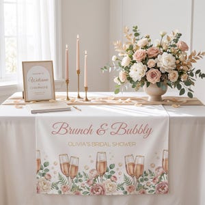 Pu&ograve; includere: Allestimento tavola per un addio al nubilato con tovaglia bianca e runner con la scritta "Brunch & Bubbly" e illustrazioni di calici di champagne. La scena comprende una composizione floreale, candele e un cartello di benvenuto, creando un'atmosfera elegante.