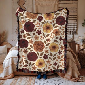 May include: A floral throw blanket with a cream background and a repeating pattern of burgundy, brown, and yellow flowers. The blanket has a black fringe border and is held up in front of a bed.
