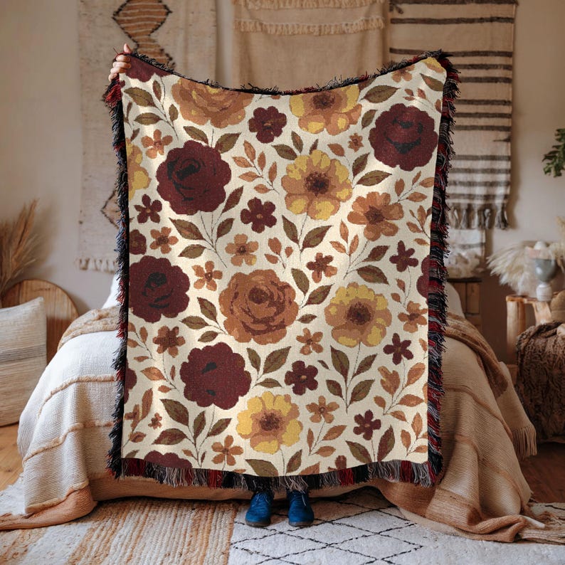 May include: A floral throw blanket with a cream background and a repeating pattern of burgundy, brown, and yellow flowers. The blanket has a black fringe border and is held up in front of a bed.