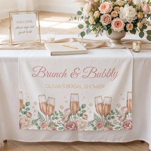 Pu&ograve; includere: Una tovaglia bianca con la scritta "Brunch & Bubbly" e "Olivia's Bridal Shower" in corsivo rosa. La tovaglia presenta un design floreale e calici di champagne. Un cartello dorato recita "Benvenuti, si prega di firmare il nostro libro degli ospiti".