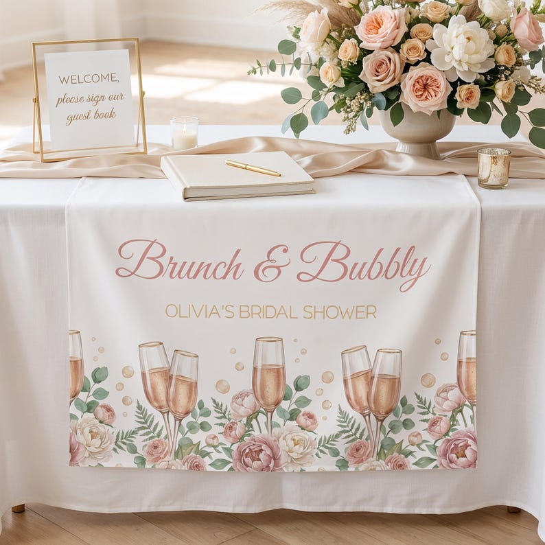 Pu&ograve; includere: Una tovaglia bianca con la scritta "Brunch & Bubbly" e "Olivia's Bridal Shower" in corsivo rosa. La tovaglia presenta un design floreale e calici di champagne. Un cartello dorato recita "Benvenuti, si prega di firmare il nostro libro degli ospiti".