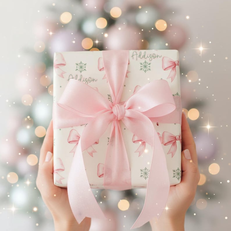 A close-up of hands holding a gift wrapped in pink bow paper against a softly lit Christmas tree backdrop with sparkling lights.