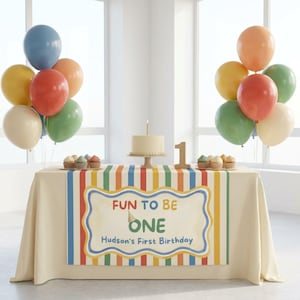 May include: A birthday table setup with a cream tablecloth featuring a colorful striped runner and a sign that reads Fun To Be One Hudson's First Birthday. The table holds a cake, cupcakes, a number one sign, and clusters of multi-colored balloons.