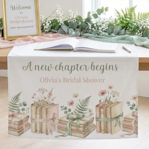 May include: A white table runner with the text "A new chapter begins" and "Olivia's Bridal Shower." The runner features watercolor illustrations of books tied with ribbons and floral accents. A sign that says "Welcome to Olivia's Bridal Shower" is in the background.