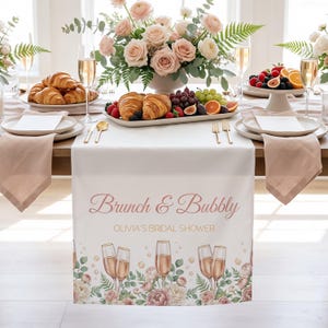 Pu&ograve; includere: Una tovaglia bianca con la scritta "Brunch & Bubbly" e "Olivia's Bridal Shower" in corsivo rosa. La tovaglia &egrave; decorata con calici di champagne e motivi floreali. La tavola &egrave; apparecchiata con piatti, tovaglioli e posate dorate, con un centrotavola di rose e croissant.