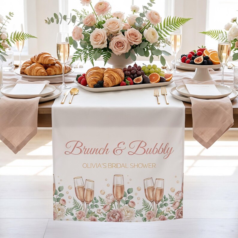 Pu&ograve; includere: Una tovaglia bianca con la scritta "Brunch & Bubbly" e "Olivia's Bridal Shower" in corsivo rosa. La tovaglia &egrave; decorata con calici di champagne e motivi floreali. La tavola &egrave; apparecchiata con piatti, tovaglioli e posate dorate, con un centrotavola di rose e croissant.