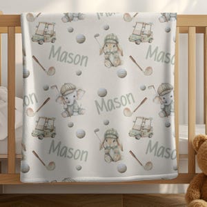 Puede incluir: Una manta para bebé de color claro con un patrón repetido de ilustraciones con temática de golf. El diseño incluye carritos de golf, pelotas de golf, palos de golf y adorables personajes de animales con atuendos de golf. El nombre "Mason" está impreso.